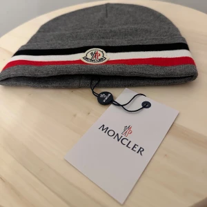 Grå Moncler mössa med ränder - Snygg grå mössa Moncler med klassiska svarta, vita och röda ränder längs kanten. Märkets logga är broderad framtill. Mössan är stickad och har en mjuk, skön känsla som passar perfekt när det är kallt ute.