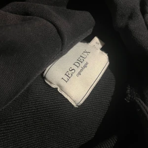 Svart hoodie från Les Deux - Svart hoodie från Les Deux med klassisk huva och snörning. Tröjan har en enkel design med vit logotyp på bröstet, ribbade muddar och en stor magficka. Perfekt för chill dagar och streetwear-stil.