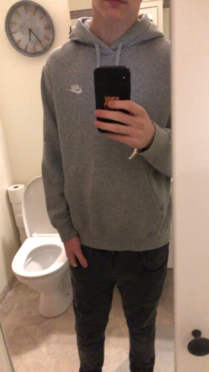 Grå Nike hoodie med huva och snörning - Grå hoodie från Nike med klassisk logga på bröstet. Tröjan har huva med snörning och en stor magficka framtill. Långärmad och i mjukt bomullsmaterial, perfekt för chill eller häng med kompisar. Enkel och stilren design som funkar till jeans eller joggers.