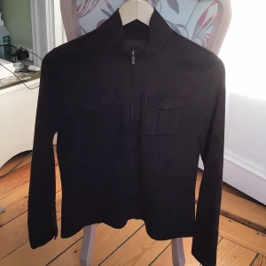 Mörkbrun mockaliknande jacka Ralph - Snygg mörkbrun jacka från Ralph 1967 i mockaliknande polyester. Jackan har raka linjer, två bröstfickor med knapp, dragkedja framtill och knappar vid ärmslut. Perfekt för dig som gillar stilren och klassisk look.