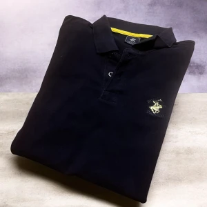 Marinblå långärmad pikétröja Beverly Hills Polo Club - Svart långärmad pikétröja från Beverly Hills Polo Club med gul broderad logga på bröstet. Klassisk krage, knappar framtill och diskreta gula detaljer vid insidan av kragen och nederkant. Tillverkad i mjuk bomull som är skön mot huden.