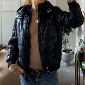Svart pufferjacka från Pepe Jeans - Svart pufferjacka från Pepe Jeans med quiltad design, huva och coola patchar på ärmen. Jackan har dragkedja framtill, ribbade muddar och snygga detaljer vid kragen.  Extremt cool men tyvärr är den för liten på mig. 