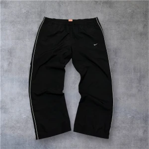 Svarta träningsbyxor från Nike XL - Svarta träningsbyxor från Nike med vita detaljer längs sidorna och klassisk Swoosh-logga på benet. Byxorna har elastisk midja och sidofickor, tillverkade i lätt polyester som andas. Perfekta för sport eller chill, loose fit och riktigt skön känsla. Skulle säga att den passar M/L. Längd 100cm.