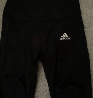 Svarta leggings från Adidas - Säljer ett par svarta leggings från Adidas med vit logga på höften. De har en tight passform och är gjorda i ett stretchigt syntetmaterial som passar perfekt för träning. Snygga och bekväma att ha på gymmet eller till vardags.