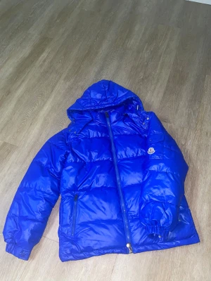 Blå Moncler pufferjacka med huva - Säljer en fet blå pufferjacka från Moncler med huva och coola detaljer som dubbla dragkedjor och logga på ärmen. Jackan har quiltad design, två fickor med dragkedja och ser riktigt varm ut. Insidan har en comic strip-etikett och DIST-märkning.
