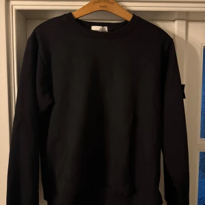 Svart sweatshirt från Stone Island - Svart crewneck sweatshirt från Stone Island med klassisk rund hals och ribbade muddar. Ikonisk patch med logga på vänster ärm. Tröjan är tillverkad i mjuk bomull och har en clean, stilren look som funkar till streetwear och chill dagar.