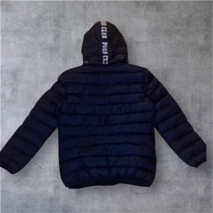 Marinblå Polo Club pufferjacka - Snygg marinblå pufferjacka från Polo Club med huva och logga på både bröstet och huvan. Jackan har dragkedja framtill, två sidofickor med dragkedja och quiltad design. Perfekt för kalla dagar och har en modern look som passar streetstyle.