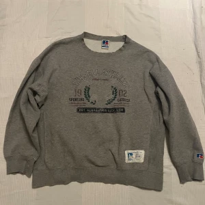 Grå sweatshirt från Russell Athletic - Grå sweatshirt från Russell Athletic med broderad logga och text framtill. Tröjan har rund hals, ribbade muddar och en patch vid nederkanten. Klassisk loose fit och långärmad, perfekt för chill dagar eller sportig stil. Sliten i ärmarna.