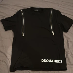 Svart DSQUARED2 t-shirt med dragkedjor - Svart t-shirt från DSQUARED2 med vita tryck fram och bak. Snygga silverfärgade dragkedjor på axlarna ger en cool detalj. Klassisk rund hals och korta ärmar. Materialet känns som mjuk bomull och passformen är normal.