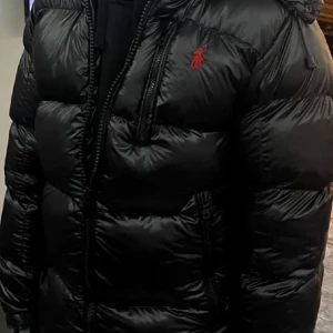 Svart pufferjacka från Polo Ralph Lauren - Svart pufferjacka från Polo Ralph Lauren med röd broderad logga på bröstet. Jackan har huva, flera dragkedjefickor och en glansig finish. Perfekt för kalla dagar och har en modern, oversized look. Materialet är syntet och dun, med långa ärmar och robust dragkedja framtill.