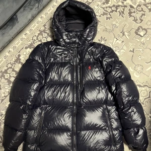 Svart pufferjacka från Polo Ralph Lauren - Svart pufferjacka från Polo Ralph Lauren med glansig finish, röd broderad logga på bröstet och hög krage med snörning. Jackan har dragkedja framtill, två sidofickor med dragkedja och en stor huva. Perfekt för kalla dagar och riktigt snygg streetstil.