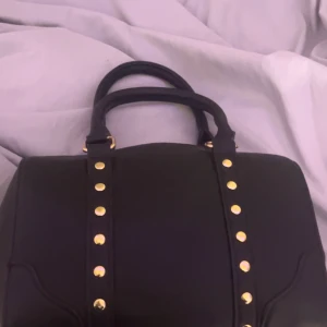 Svart handväska med gulddetaljer - Snygg svart handväska i skinn med guldfärgade nitar och detaljer. Väskan har två stadiga handtag och en dragkedja upptill. Stilren och klassisk form som passar till många olika outfits. Perfekt storlek för att få plats med allt viktigt.