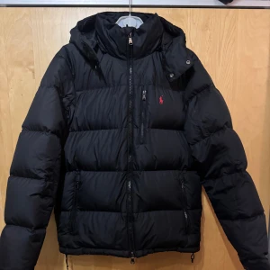 Svart pufferjacka från Polo Ralph Lauren - Svart pufferjacka från Polo Ralph Lauren med huva och dragkedja framtill. Jackan har ett rött Polo-logo broderat på bröstet, flera fickor med dragkedja och en stilren design. Perfekt för kalla dagar och har en modern, clean look.