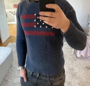 Lexington mörkblå stickad tröja  - Snygg mörkblå stickad tröja från Lexington med amerikansk flagga i rött och vitt på bröstet. Tröjan har rund hals, ribbade muddar och lång ärm. Perfekt för dig som gillar klassisk stil med en twist.