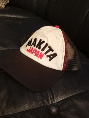 Makita Japan keps i svart och vitt - Snygg keps med Makita Japan-tryck framtill. Framsidan är vit med svart och röd text, skärmen är svart och baksidan har svart mesh med röda detaljer. Perfekt för dig som gillar streetstyle och vill sticka ut med ett coolt varumärke.