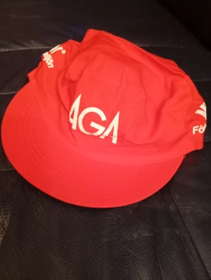 Röd AGA keps med tryck - Säljer en röd keps med vit AGA-logga och texten 'Förbättra arbetsmiljön' tryckt på sidan. Kepsen har klassisk form med böjd skärm och är tillverkad i lätt bomullstyg. Perfekt för dig som gillar retro eller vill sticka ut med en unik accessoar.