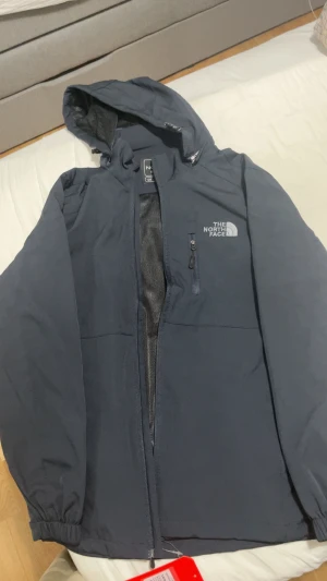 Mörk blå vindjacka The North Face - Säljer en mörkblå vindjacka från The North Face med huva och dragkedja framtill. Jackan har en bröstficka med dragkedja, elastiska muddar och justerbar nederkant. Perfekt för blåsiga dagar och enkel att matcha med streetwear. Storleken är M men på jackan står det XL