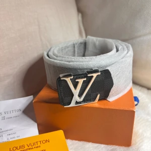 Louis Vuitton rutig svart bälte - Snyggt svart bälte från Louis Vuitton med klassiskt rutigt mönster och en stor LV-spänne i silver. Bältet är tillverkat i skinn och har en stilren, modern look som passar perfekt till jeans eller kostymbyxor.
