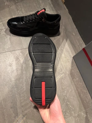 Svarta sneakers från Prada - Snygga svarta sneakers från Prada med glansiga detaljer och meshpaneler. Skorna har rund tå, platt sula och klassisk snörning. Röd Prada-logga syns på plösen och under sulan. Perfekta för dig som gillar stilrena och exklusiva sneakers.