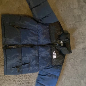 Blå och svart pufferjacka The North Face - Säljer en blå och svart pufferjacka från The North Face med broderad logga på bröstet och ryggen. Jackan har hög krage, dragkedja framtill och två fickor med dragkedja. Perfekt för kalla dagar och riktigt snygg streetstil.
