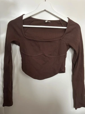 Brun croppad långärmad topp - Säljer en brun croppad topp med lång ärm och fyrkantig halsringning. Toppen är ribbad och har en snygg sömdetalj över bröstet som ger en cool look. Perfekt att styla med höga jeans eller kjol för en trendig vibe. Lite korsett vibe