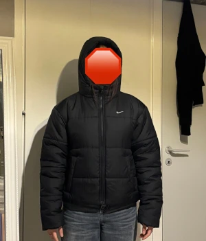 Svart pufferjacka från Nike - Säljer en svart pufferjacka från Nike med huva och dragkedja framtill. Jackan har en boxig passform, långa ärmar och diskret Nike-logga på bröstet. Perfekt för kalla dagar och riktigt snygg till jeans eller cargo. Materialet är syntet och jackan är quiltad för extra värme.
