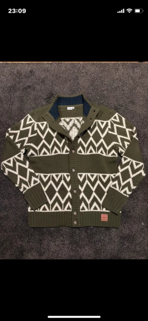 Grön kofta med vitt zigzag-mönster - Snygg grön kofta från Junkyard med vitt zigzag-mönster. Tröjan har ribbade muddar, knappar framtill och en mörkblå insida vid kragen. Stickad i mjukt material och perfekt för kyliga dagar. Passar dig som gillar grafiska mönster och vill ha en unik look.