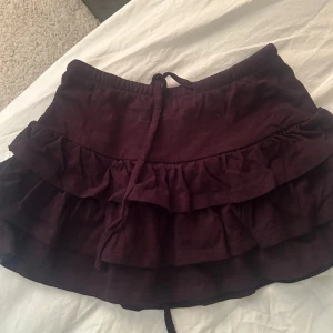 Svart volangkjol från Gina Tricot - Supersöt mörklila kortkjol från Gina Tricot med dubbla volanger och knytband i midjan. Kjolen har en skön passform och är perfekt för dig som gillar en feminin och trendig stil. Tillverkad i mjukt bomullstyg.