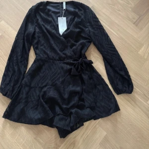 Svart omlottklänning med knyt - Supersnygg svart kortklänning från NA-KD med omlottmodell och v-ringning. Klänningen har långa ärmar, ett coolt strukturerat mönster och ett knyt i midjan. Materialet är lätt och transparent med vågiga detaljer som ger en unik look.