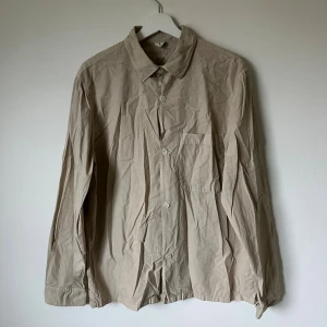 Beige överskjorta från ARKET, regular fit - Snygg beige överskjorta från ARKET i regular fit. Skjortan har klassisk krage, bröstficka och knäppning framtill med vita knappar. Tillverkad i mjuk bomull, perfekt för en clean och chill look. Långärmad och enkel design som funkar till många stilar. 5J9