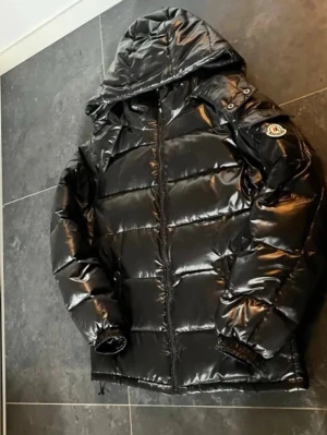 Svart pufferjacka med huva Fashion - Säljer en svart, glansig pufferjacka från Fashion med huva och dragkedja framtill. Jackan har ett tydligt märke på ärmen och elastiska muddar vid ärmsluten. Perfekt för kalla dagar och riktigt snygg till streetwear-looken.