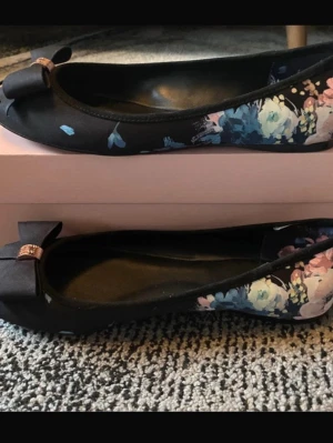 Pretty ballerinas Svarta med blommigt mönster - Snygga svarta ballerinaskor från Pretty Ballerinas i storlek 39. Skorna har ett blommigt mönster i blått, rosa och vitt på sidorna samt en dekorativ rosett framtill med gulddetalj. Mjukt material och rund tå gör dem både bekväma och stilrena. Aldrig använda, bara provat dem inomhus 1 gång. 