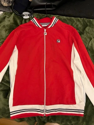 Röd och vit vintage jacka från Fila - Säljer en snygg vintage röd vindjacka från Fila med vita paneler längs sidorna och ärmarna. Denna säljs ej längre och är i väldigt bra skick! Den har en liten defekt (se sista bilden) på sidan av jackan, men inget som läggs märke till!