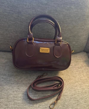 Vinröd handväska Lady's Collection - Snygg mörkbrun handväska från Lady's Collection i glansigt skinn med guldfärgade metalldetaljer. Väskan har rundade handtag och avtagbar axelrem, perfekt för att bära både i handen och över axeln. Stilren design med metallplakett framtill.