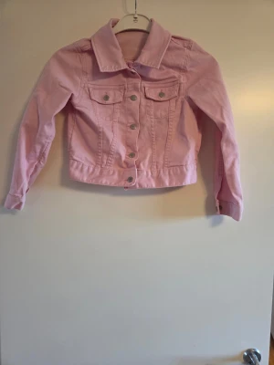 Ljusrosa jeansjacka med knappar, stl 128 - Snygg ljusrosa jeansjacka från H&M med klassisk krage, silverfärgade knappar och två bröstfickor med lock.
