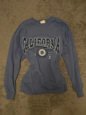 Blå sweatshirt - Säljer en blå sweatshirt från Pull & Bear med California-tryck på bröstet. Säljer då den inte kommit till användning. Har använt den max 3 gånger, därav priset.