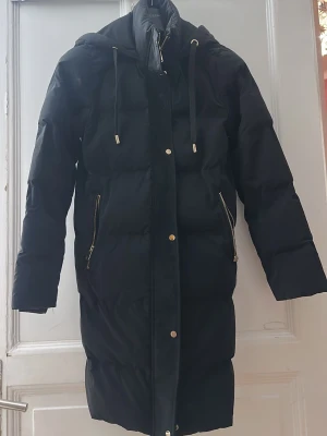Svart lång pufferjacka Zara - Svart pufferjacka från Zara med huva och långa ärmar. Jackan har dragkedja och knappar framtill, samt två sidofickor med dragkedja. Lång modell som ger extra värme och stil, perfekt för kalla dagar. Materialet är polyester.