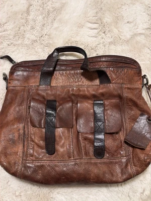 Brun skinnportfölj med fickor - Säljer en klassisk portfölj i brunt skinn med två framfickor och mörkare bruna detaljer. Väskan har både handtag och axelrem, samt flera fack och dragkedjor för smart förvaring. Perfekt för laptop, böcker och allt du behöver till plugget eller jobbet.