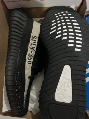 Adidas Yeezy Boost 350 V2 Svart/Vit - Säljer ett par Adidas Yeezy Boost 350 V2 i svart och vit. Skorna har en stickad ovandel med en vit rand och texten 'SPLY-350' på sidan. Sulan är tjock och ribbad med Boost-teknologi för extra komfort. Perfekt för dig som gillar streetwear och vill ha något unikt.