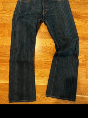 Mörkblå Nudie Jeans med breda ben - Säljer ett par mörkblå jeans från Nudie med klassisk orange söm och snygga broderade bakfickor. Jeansen har breda ben och är gjorda i slitstark jeansmaterial med selvedge-detalj vid benslutet. Perfekt för dig som gillar loose fit och streetstil. Mått: midja 39cm, längd 105cm, benöppning 22cm, lår 27 cm