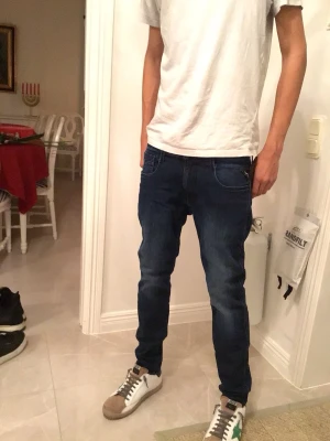 Replay Anbass mörkblå jeans - Säljer ett par Replay Anbass jeans i mörkblå tvätt med snygga slitningar. Modellen har smal passform, klassiska fem fickor och dragkedja med knapp. Materialet är stretchigt jeans och byxorna har Replay-logga på fickan. Perfekt för dig som gillar stilrena och moderna jeans.