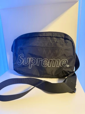 SUPREME Shoulderbag/Axelväska - Detta är en supreme(FW18)/2018 axelväska med 3 olika utrymmen, 2 med dragkedja 1 med kardborre, justerbart axelband och möjlighet till olika lösningar med 2 kardborre-band på baksidan.