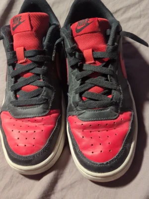 Nike svart och röd sneakers - Säljer ett par Nike sneakers i svart och rött med klassisk design. Skorna har snörning, perforerad tå och vit sula. Ovandelen är i skinn och mesh, vilket ger både stil och komfort. Perfekt för dig som gillar streetwear och ikoniska sneakers.