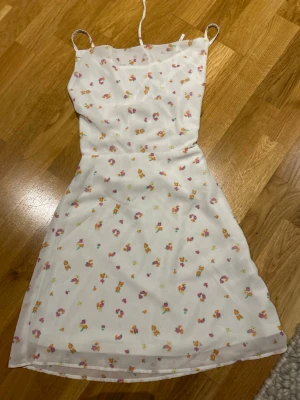 Vit blommig kortklänning  - Superfin vit klänning med små färgglada blommor och smala axelband. Klänningen har snörning i ryggen och är lätt utsvängd nertill. Perfekt för sommarens alla tillfällen, med ett luftigt och mjukt material som känns skönt mot huden.
