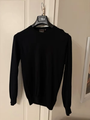 Svart stickad tröja Oscar Jacobson - Svart stickad tröja från Oscar Jacobson, tillverkad i Italien. Klassisk rund hals och långärmad modell med ribbade muddar vid ärmslut och nederkant. Perfekt för dig som gillar stilrena och enkla plagg med premiumkänsla.