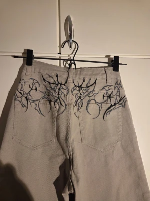  Tribaltryck custom en i hela världen jeans  - Unika beige jeans med breda ben och handmålat tribaltryck i svart och vitt på både framsidan och baksidan. Coola detaljer med japanska och koreanska tecken på vänster ben. Materialet är kraftig bomull och modellen är högmidjad med fem fickor.