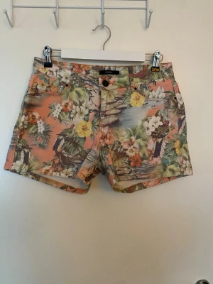 Coola denimshorts från Lindex - Snygga shorts från Lindex i denim med tropiskt blommönster i orange, grönt, gult och vitt. Klassisk femficksmodell med knapp och dragkedja framtill. Perfekta för varma dagar när du vill sticka ut lite extra.