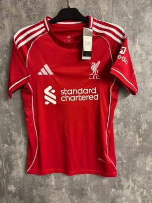 Liverpool Salah - Ny Storlek S - Bilden visar en röd Liverpool FC fotbollströja med M. Salah och nummer 11 på ryggen, tillverkad av Adidas. Tröjan har vita detaljer och tryck, samt klubbens logga på bröstet.