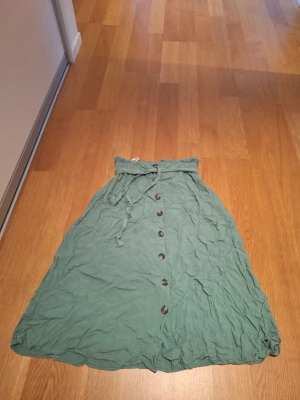 Grön långkjol med knappar från Pieces - Säljer en ljusgrön långkjol från Pieces i storlek XS. Kjolen har knappar hela vägen framtill och ett knytband i midjan. Perfekt för dig som gillar en avslappnad och trendig stil. Materialet känns lätt och flowy.