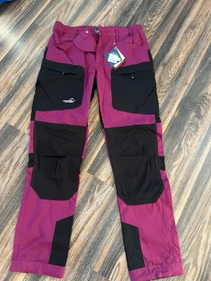 Fuchsia och svarta friluftsbyxor Arrak Outdoor - Säljer ett par snygga fuchsia och svarta friluftsbyxor från Arrak Outdoor, modell Active Stretch Pants LADY. Byxorna har flera praktiska fickor med dragkedja, förstärkta partier och är gjorda i slitstarkt syntetmaterial. Perfekta för utomhusaktiviteter och äventyr. Aldrig använda!